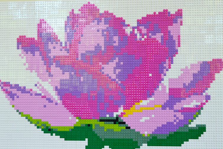 A detailed LEGO mosaic displaying a bright pink lotus flower, with purple shadows and pastel highlights with a green stamen against a stark white background.