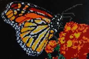 A LEGO mosaic by artist Brian Korte, depicting a vivid orange monarch butterfly pollinating red and orange flowers.