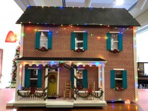 Exterior view of a two-story brick dollhouse with teal shutters, decorated for the holidays with colored lights