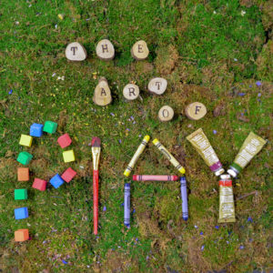 Wooden letters spell out "the art of" and various art items spell out "play." The words are sitting on top of a moss background.