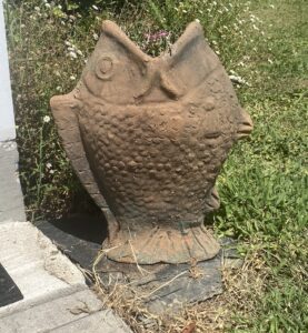A concrete, fish shaped potting container sitting on the grass by a small slab of pavement.