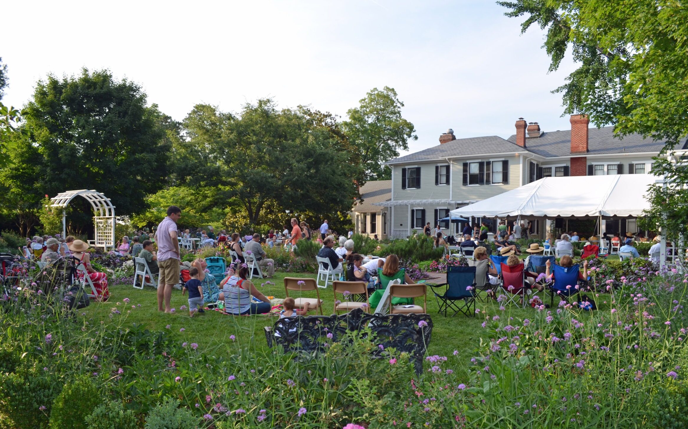 An image of a concert happening outside Bloemendaal House at Lewis Ginter Botanical Garden. People are sitting in the grass, walking around, eating, drinking, and enjoying the music.